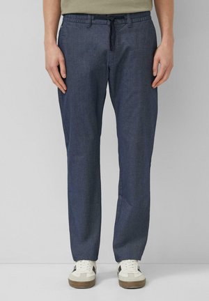 Persoon draagt blauwe vrijetijdsbroek met aantrekkoord in de taille en witte sneakers met zwarte accenten, staand tegen een effen achtergrond.