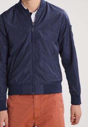 Homme portant une veste bomber bleu marine zippée par-dessus une chemise blanche avec un pantalon orange, debout devant un fond uni.