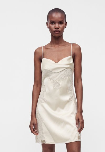 Femme aux cheveux courts portant une robe nuisette en satin crème avec des détails en dentelle, debout devant un fond clair uni.