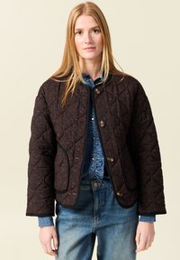 Kurtka pikowana w ciemnym brązie z subtelnym wzorem w panterkę. Posiada okrągły dekolt, zapięcie na guziki oraz kontrastowe czarne wykończenie.