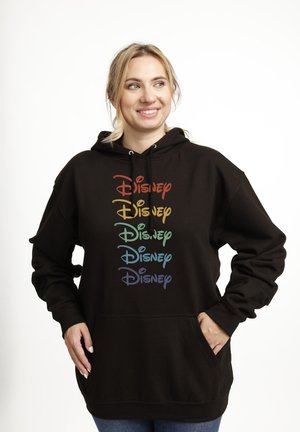 Uśmiechnięta kobieta o blond włosach, ubrana w czarną bluzę z kapturem z napisem „Disney” w pięciu tęczowych rzędach, ręce w kieszeniach, patrząca w prawo.