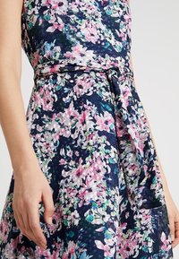 Blommig midiklänning i marinblått med rosa, gröna och vita blommor. Har ett knytbälte i midjan och en strukturerad yta.