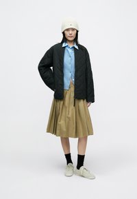 Chaqueta negra acolchada, camisa azul claro abotonada, falda plisada caqui, calcetines negros y zapatillas claras. Gorro blanco de punto con logo.