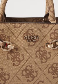 Ruskean kuvioitu käsilaukku, jossa on lomittain olevat 'G'-kuviot, kultaiset metalliosat ja yläkahvat. Materiaali näyttää teksturoidulta ja kestävältä.