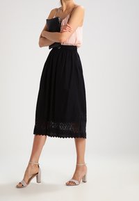 Jupe midi noire avec bordure en dentelle à l'ourlet, associée à un haut rose clair. La mannequin porte des sandales à talons carrés beige et tient une pochette noire.