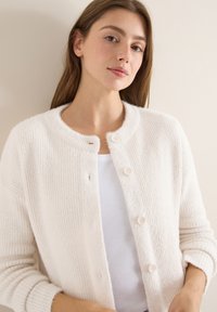 Weißer gerippter Cardigan mit Rundhalsausschnitt, fünf Knöpfen und langen Ärmeln. Aus weichem, strukturiertem Stoff gefertigt. Über einem weißen Oberteil getragen.