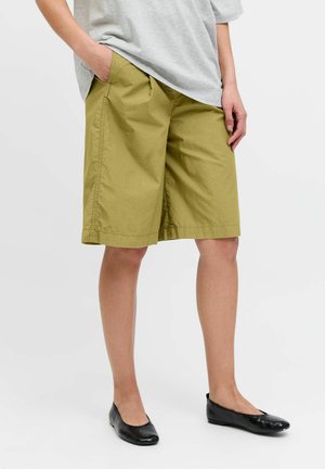 Person som har på seg knelange olivengrønne shorts, grå t-skjorte og svarte ballettsko, står mot en hvit bakgrunn.