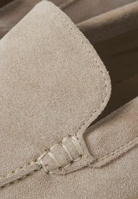 Detailní záběr béžových semišových mokasín ukazujících detailní šití a strukturovaný svrchní materiál.