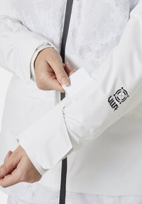 Hvit vanntett jakke med teksturert overflate, en svart logo på ermet, og en hånd som setter et hvitt kort i en lomme.