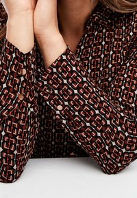 Hnědo-bílá blůza se vzorem s geometrickými tvary, dlouhé rukávy s viditelnými detaily knoflíků a hladkým textilním povrchem.