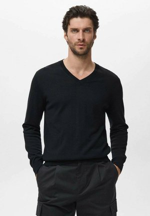 Hombre con cabello corto rizado y barba, vestido con un suéter negro de cuello en V y pantalones negros, con las manos en los bolsillos frente a un fondo liso.