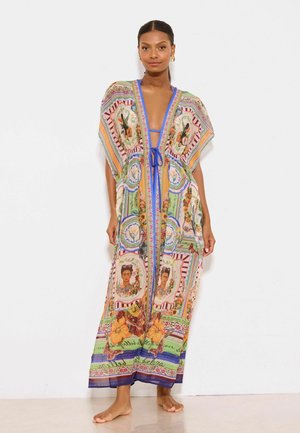 Frau steht barfuß und trägt einen bunten, gemusterten langen Kaftan mit floralen und Portrait-Designs über einem blauen Bikini.