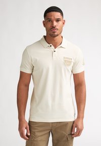 Beige puuvillapikéepaita, jossa on klassinen kaulus, kolmen napin läppä ja rintatasku, jossa on logoplässi.