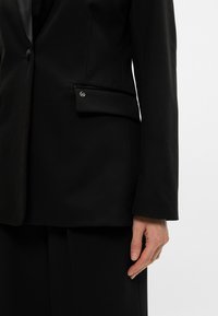 Černý blazer s jedním knoflíkem, s klopami a boční kapsou. Hladká látka s přiléhavým střihem a čistými liniemi.