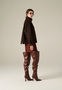 Giacca oversized marrone di lana con colletto alto, abbinata a una mini gonna aderente color ruggine e stivali alti fino al ginocchio in pelle lucida marrone.