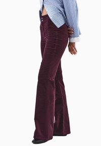 Burgundowe spodnie kordurowe z szerokimi nogawkami, z kieszeniami z przodu i metalowymi guzikami, noszone z niebiesko-białą w paski koszulą, pokazane z boku.