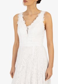 Robe blanche en dentelle avec un décolleté en V profond, motifs floraux, délicates bordures en dentelle aux épaules et taille cintrée, s'évasant en une jupe pleine.