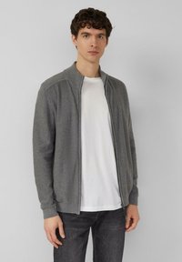 Grijze cardigan met ritssluiting, geribbelde kraag en manchetten, lichtgewicht gebreide textuur, gedragen over een wit T-shirt, gecombineerd met donkere spijkerbroek.