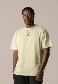 T-shirt en coton vert clair avec une coupe décontractée, des manches courtes et un col rond. Présente une broderie subtile sur la poitrine et est accessoirisé avec des colliers en or.