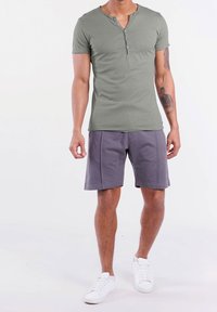 Olijfgroen T-shirt met korte mouwen, een ronde hals en drie knoopdetails, gecombineerd met grijze shorts en witte sneakers.
