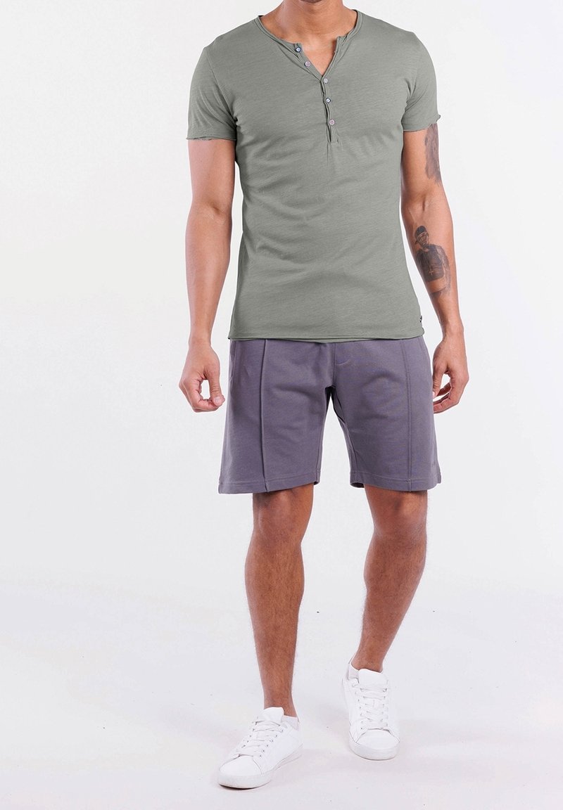 Olijfgroen T-shirt met korte mouwen, een ronde hals en drie knoopdetails, gecombineerd met grijze shorts en witte sneakers.