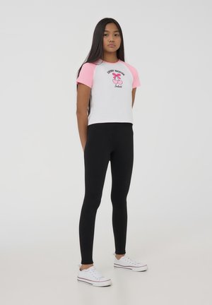 T-shirt bianca e rosa a maniche corte con grafica di ciliegia e testi, abbinata a leggings neri e sneakers bianche.