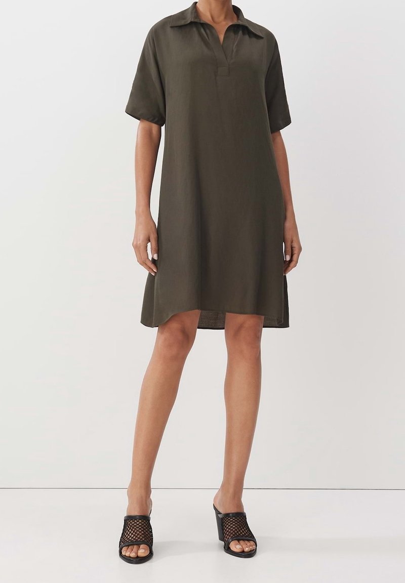 Robe chemise courte à manches courtes de couleur vert olive, avec col et silhouette droite. Portée avec des sandales noires à talons texturées. Design simple et minimaliste.