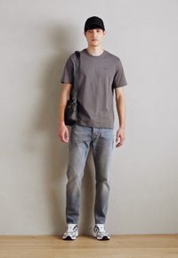 T-shirt gris à manches courtes, jeans bleu clair, casquette noire et sac à bandoulière sombre. Baskets avec des accents blancs et noirs. Fond uni.