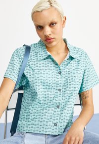 Cămașă cu mânecă scurtă, cu nasturi, dintr-un material teal deschis, având un model geometric, guler și două butoane gri. Asortată cu blugi de culoare albastru deschis.
