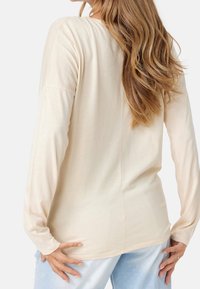 Ljusbeige långärmad topp med avslappnad passform, mjuk textur och rak nederkant. På baksidan finns en subtil sömdetalj. Matchas med ljusblå jeans.
