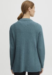 Teal stickad tröja med en strukturerad yta, öppen rygg och ribbad nederkant. Har långa ärmar och en avslappnad passform.