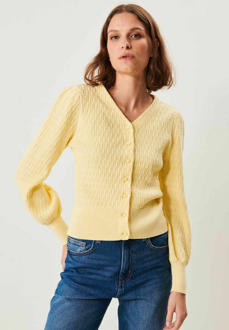 Kvinde iført en struktureret, knaplukket gul cardigan med lange ærmer og blå jeans, stående mod en ensartet lys baggrund.