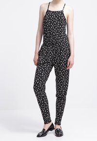 Mono negro con un patrón paisley, que presenta tirantes finos, una cintura elástica y piernas acampanadas. El material parece ligero y texturizado.