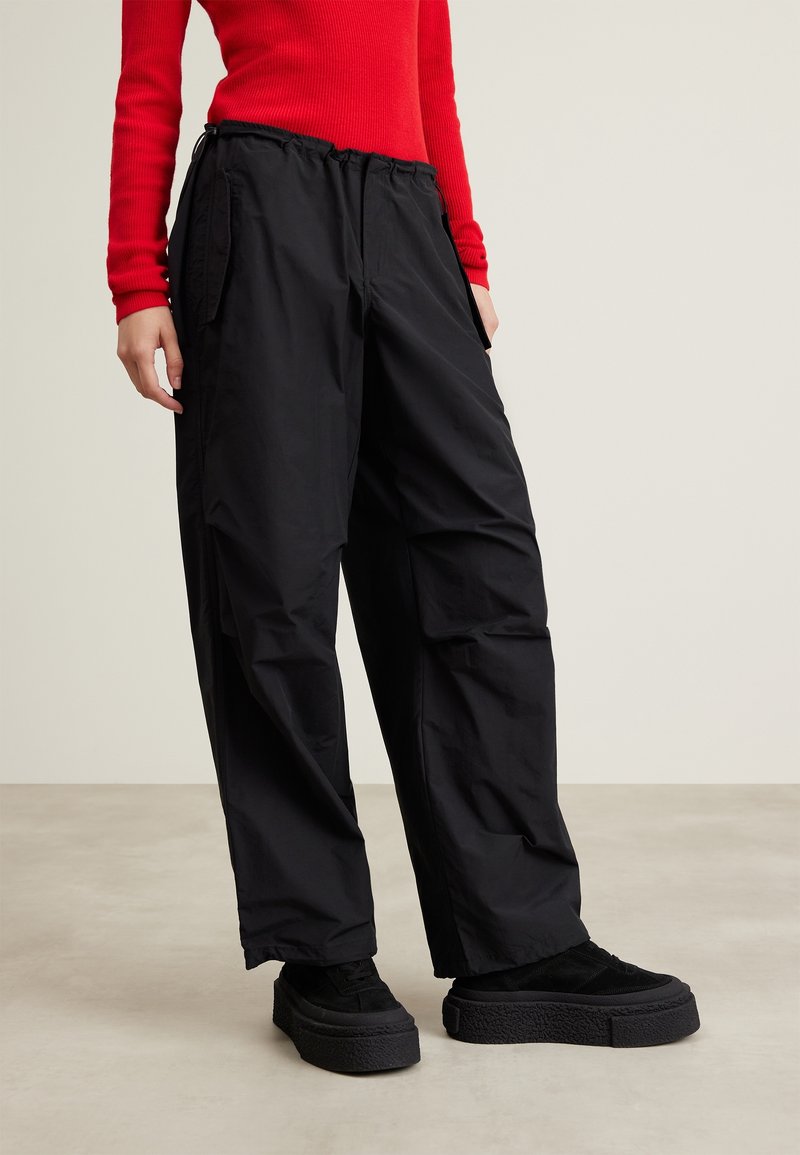 Persona che indossa pantaloni neri larghi, scarpe con tacco alto nere e un maglione a maniche lunghe rosso a costine su uno sfondo chiaro e neutro.
