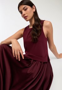 Burgundowa bluzka bez rękawów z okrągłym dekoltem, w zestawie z lekko opadającą, plisowaną spódnicą w tym samym kolorze. Gładka, satynowa tekstura.