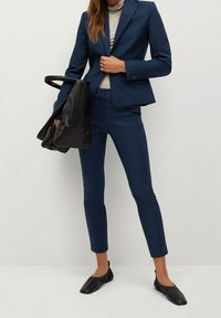 Blazer ajusté bleu marine et pantalon slim, associés à un col roulé gris clair et des chaussures plates noires. Comprend un sac fourre-tout noir.