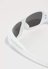 Óculos de sol brancos da Oakley com lentes cinzas, apresentando um design elegante, textura suave e um logótipo dourado na haste.