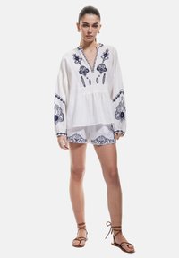 Hvid bomuldsbluse med marineblå broderi, V-hals, lange ærmer. Kombineret med matchende shorts med lignende mønstre. Brune sandaler.