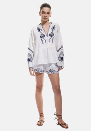 Hvid bomuldsbluse med marineblå broderi, V-hals, lange ærmer. Kombineret med matchende shorts med lignende mønstre. Brune sandaler.