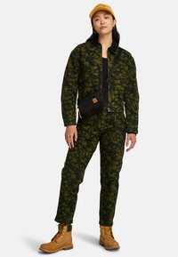 Donkergroen utility jumpsuit met een bloemen camouflagepatroon, zwarte schoudertas, tan laarzen en een gele baseballpet.