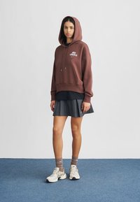 Καφέ φούτερ με κουκούλα και λογότυπο, φορεμένο πάνω από μαύρο τοπ, σετ με γκρι φουσκωτή φούστα, μπεζ αθλητικά παπούτσια και γκρι κάλτσες.