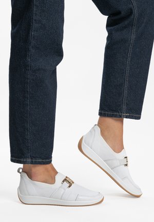 Füße in weißen Slip-On-Turnschuhen mit goldenen Schnallen und Gummisohlen, kombiniert mit dunkelblauen Jeans.
