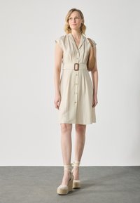 Kort, kremfarget skjortekjole med cap sleeves, belted midje og teksturert stoff; parret med høye plattform-espadrillos og en brun skulderveske.