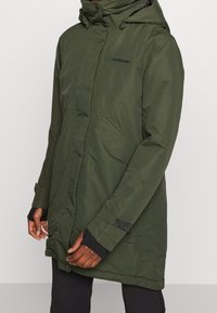 Parka impermeabile verde con chiusura a zip e a clip, cappuccio e tasche laterali. Presenta polsini neri e un logo del marchio sottile sul petto.