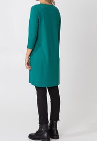 Groene tuniek met lange mouwen en een ronde halslijn, losse pasvorm en zijzakken, gecombineerd met nauwsluitende zwarte broeken en zwarte laarzen.