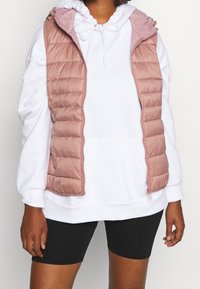Gilet rembourré rose clair avec un motif matelassé horizontal, fermeture éclair à l'avant et capuche, superposé sur un sweat à capuche blanc et un short noir.