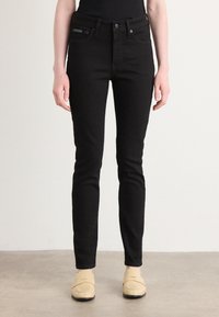Zwarte slim-fit jeans met een gladde textuur, voorzien van een klassieke tailleband, voorzakken en een knoopsluiting. Gecombineerd met lichtgekleurde schoenen.