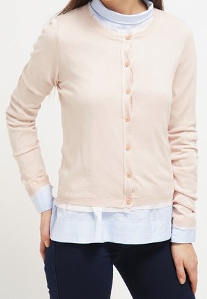 Vrouw draagt een lichtroze vest met knopen over een wit shirt en marineblauwe broek, staat met één hand op haar heup.