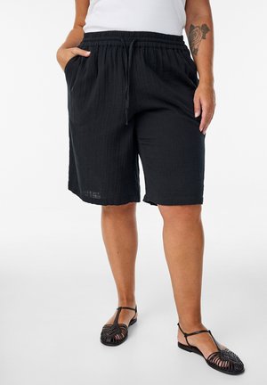 Person wearing loose black knee-length shorts, white top, black woven sandals, with rose tattoo on left forearm, hands in pockets.