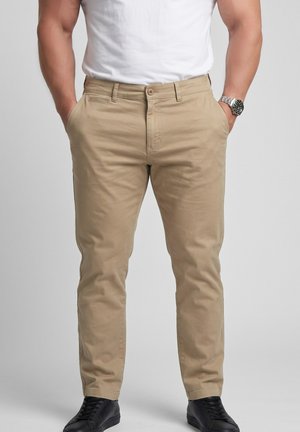 Mand iført beige chinos med lommer, en hvid t-shirt, sorte sneakers og et metal armbåndsur, stående mod en ensfarvet baggrund.